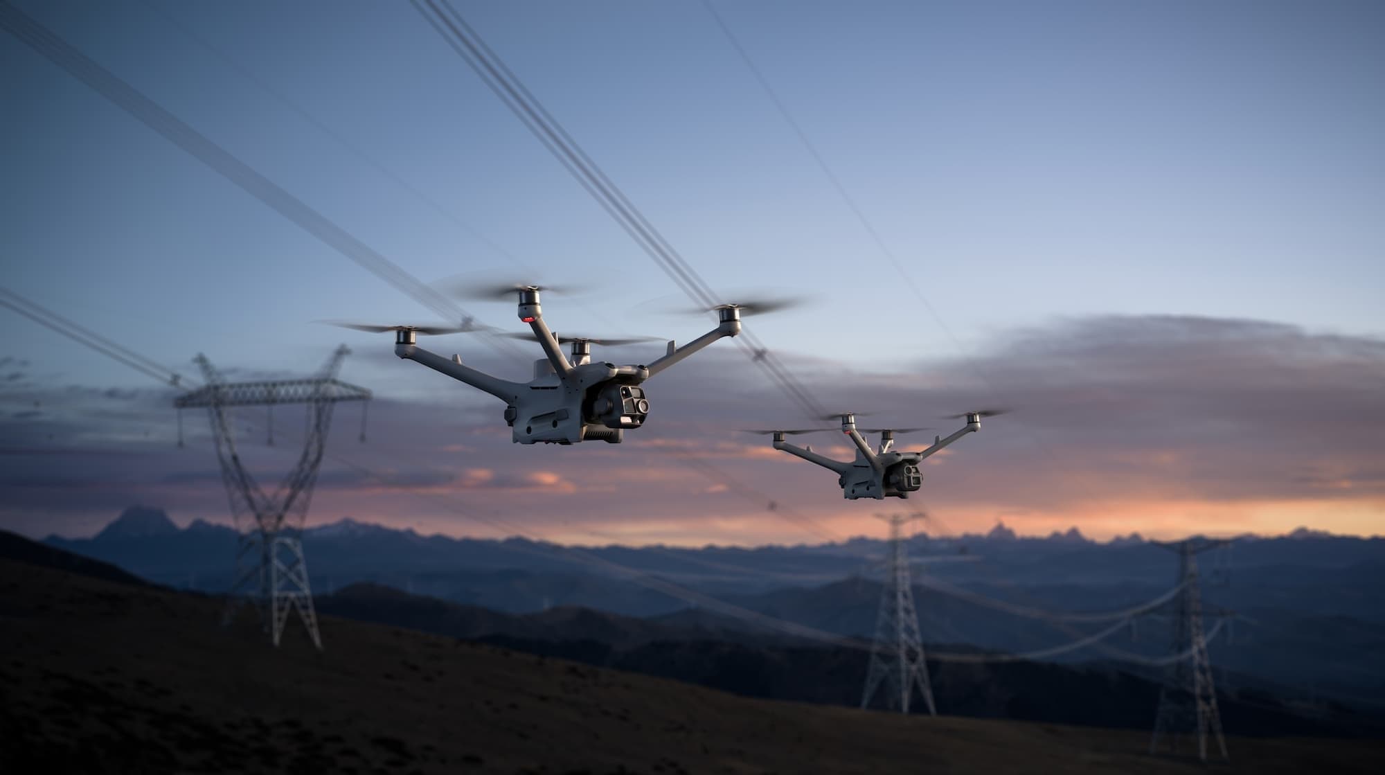 Deux drones en vol au crépuscule, au-dessus d'une ligne électrique.