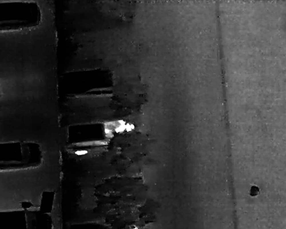 Vue thermique prise par drone, de nuit, au-dessus d'un parking : des voitures en sombre, et une personne qui ressort en blanc contre l'une d'elles.