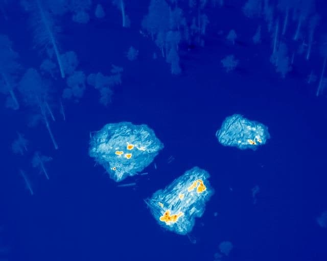 Vue thermique prise par drone au-dessus d'une forêt : trois tas de branches en feu, leurs points chauds en orange.