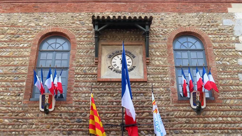 Façade en galets et briques d'un hôtel de ville orné de drapeaux français, catalans et d'une horloge
