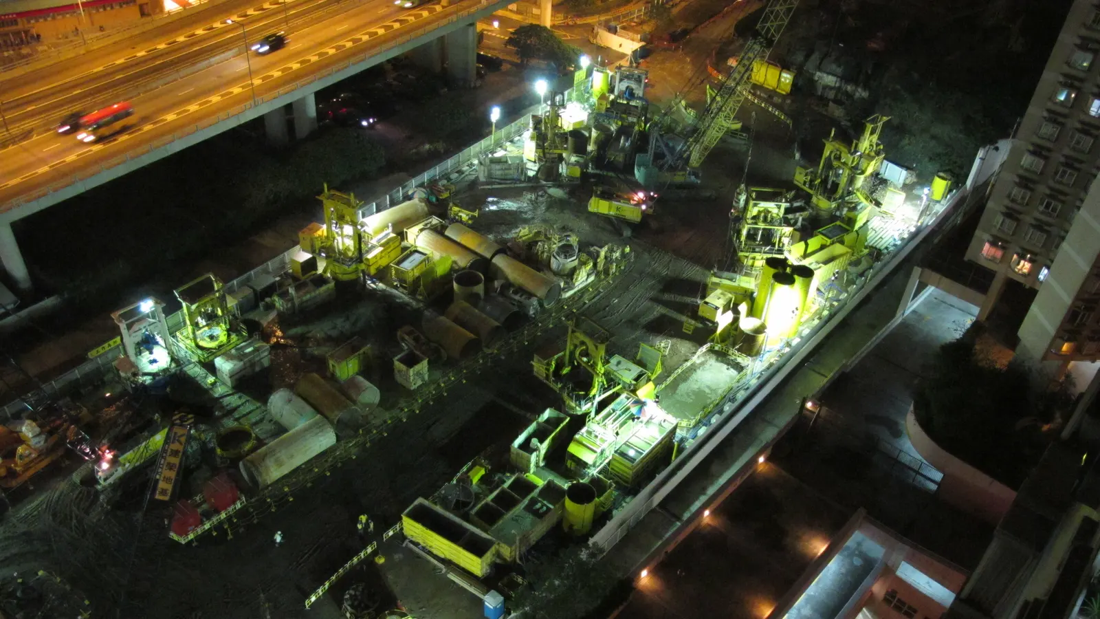 Vue aérienne de nuit d'un chantier éclairé avec engins et matériel, en contrebas d'une voie routière